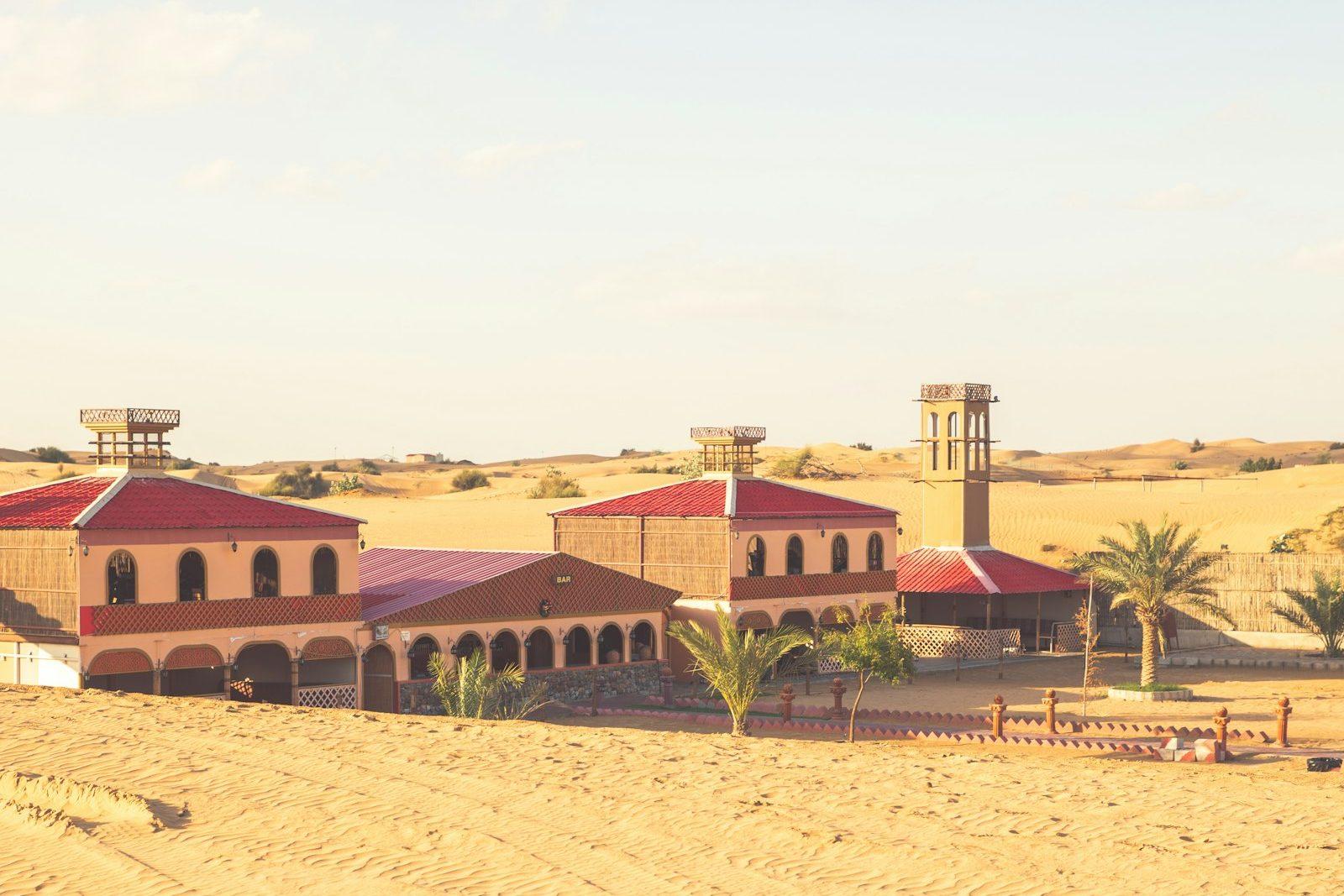 Low-rise Mediterranean-style buildings with red tile roofs and a wind tower standing at the edge of open desert sand.