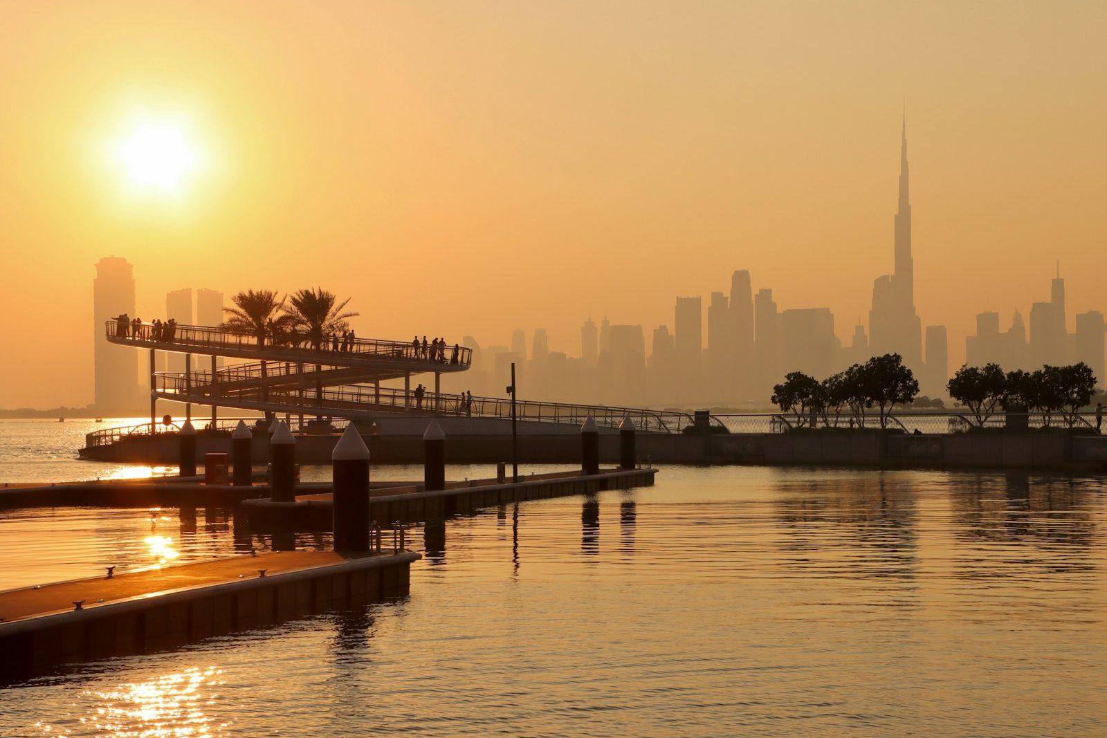 Sunset over Dubai Creek from the Creek Harbour waterfront, a timber boardwalk in the foreground and the Downtown skyline silhouetted across the water.
