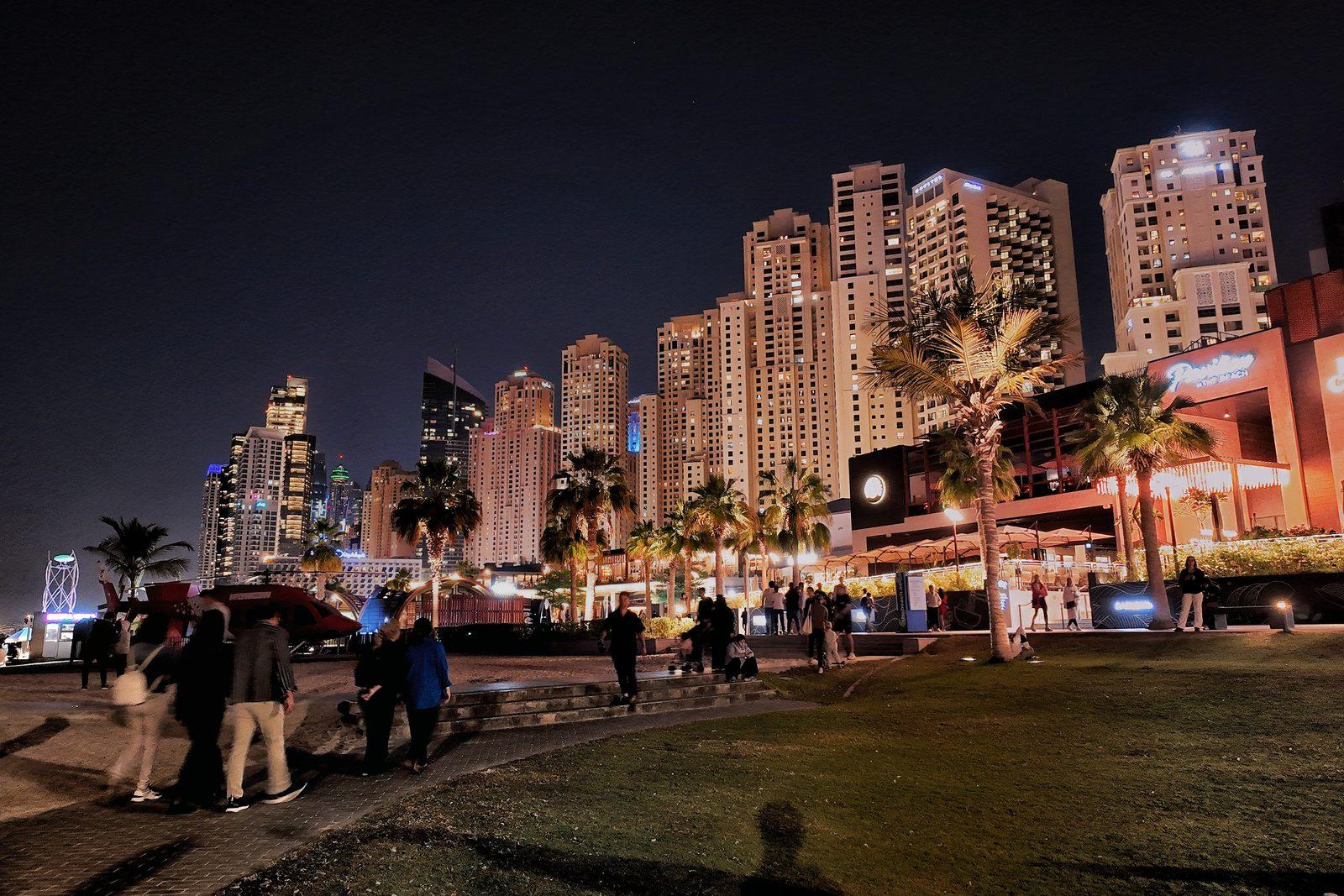 The Jumeirah Beach Residence promenade after dark, the lit wall of residential towers rising behind palm trees as people walk the waterfront.