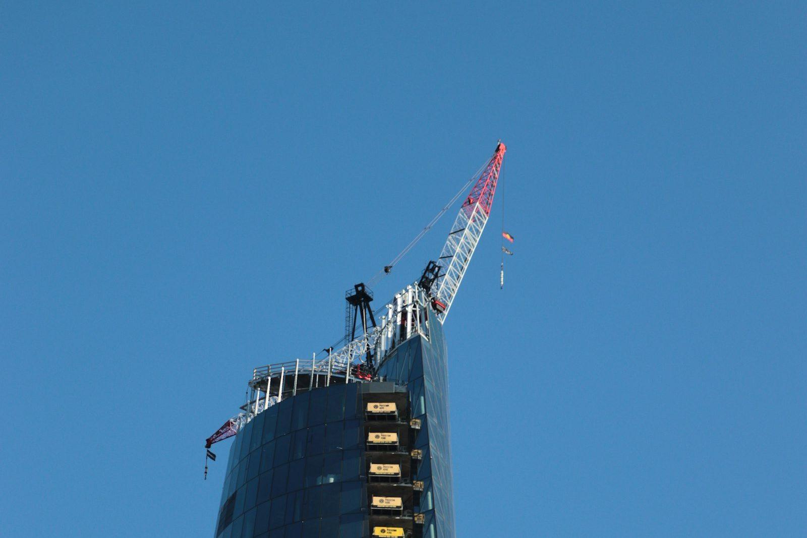 A tower still under construction against a clear blue sky, a tower crane standing on its unfinished top floors.
