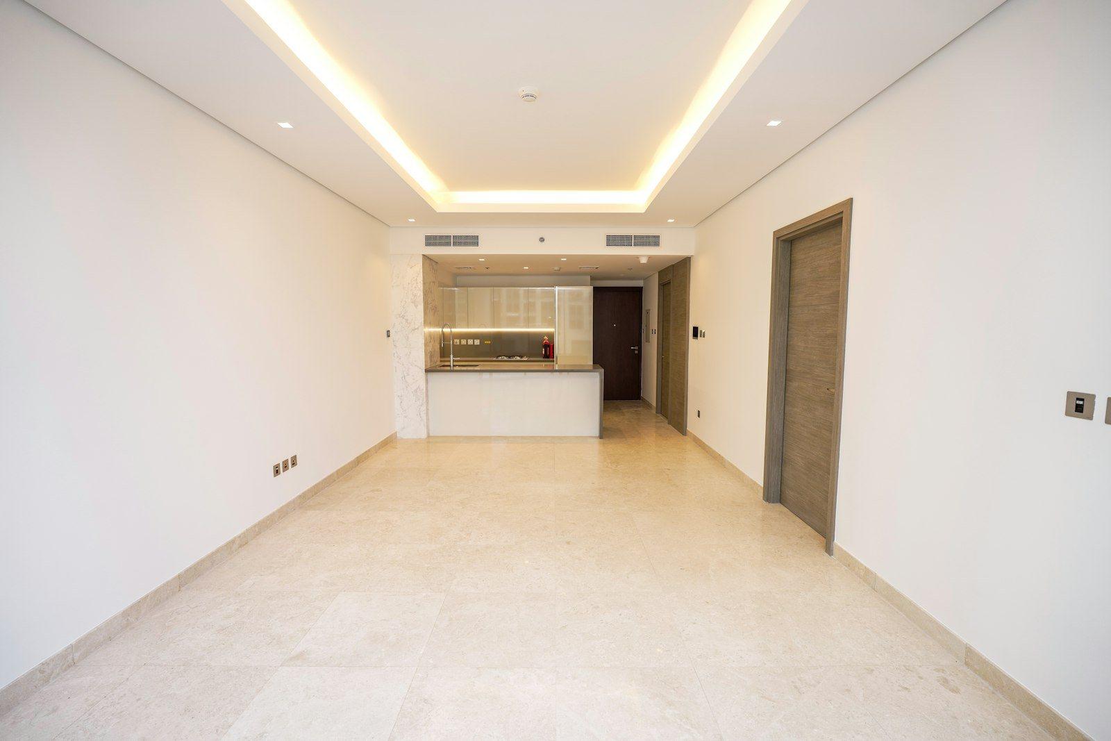 Open-plan living and dining room finished in cream marble with recessed cove lighting, a compact kitchen island in gloss white and a marble-clad return at the far end.
