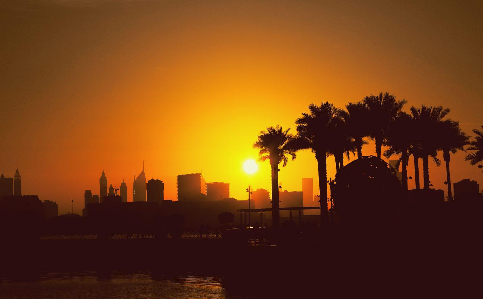 Sunset over the water in Dubai, the low sun behind a silhouetted skyline with date palms and a sphere sculpture in the foreground and the tide catching the last orange light.