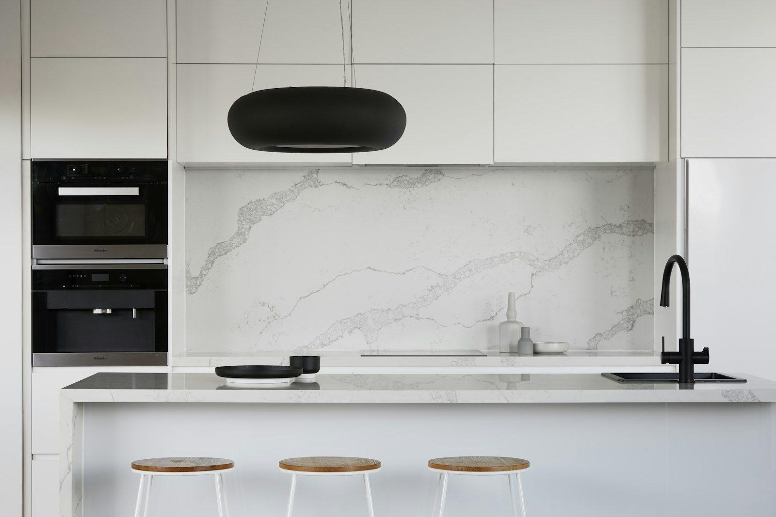 Chef’s kitchen in white with a marble-veined splashback and island top, integrated oven and steam oven, a matt black mixer tap and three oak-topped stools drawn up to the island.