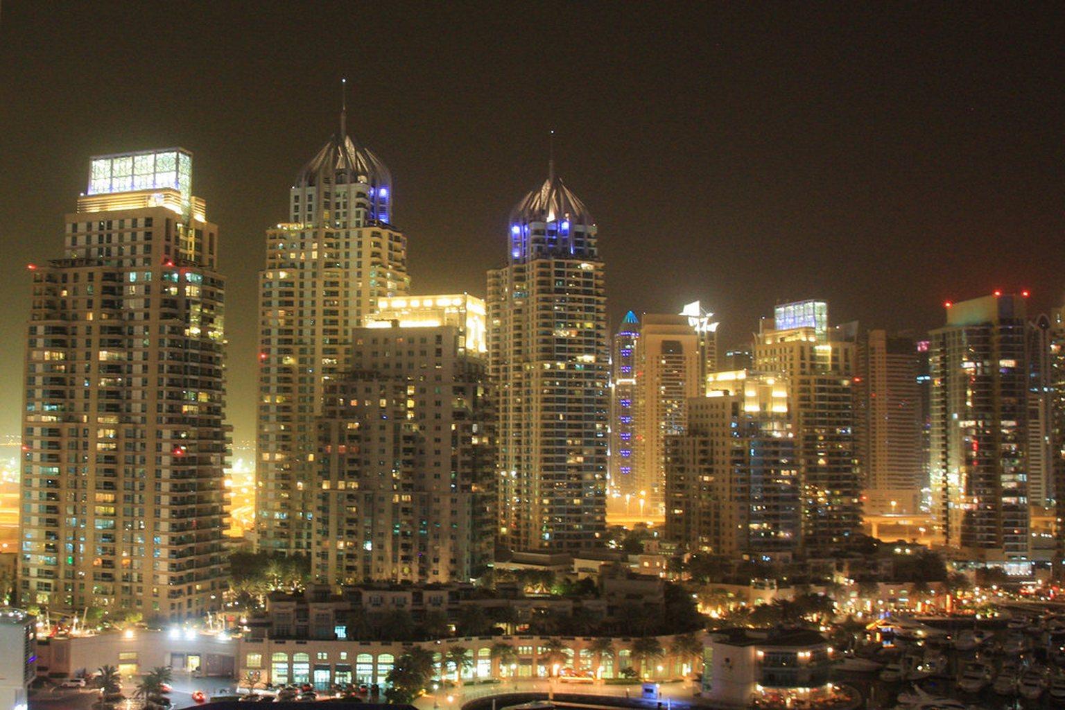 Dubai Marina towers at night viewed from a high floor, lit facades and crown lighting massed above the marina with the low-rise podium below.