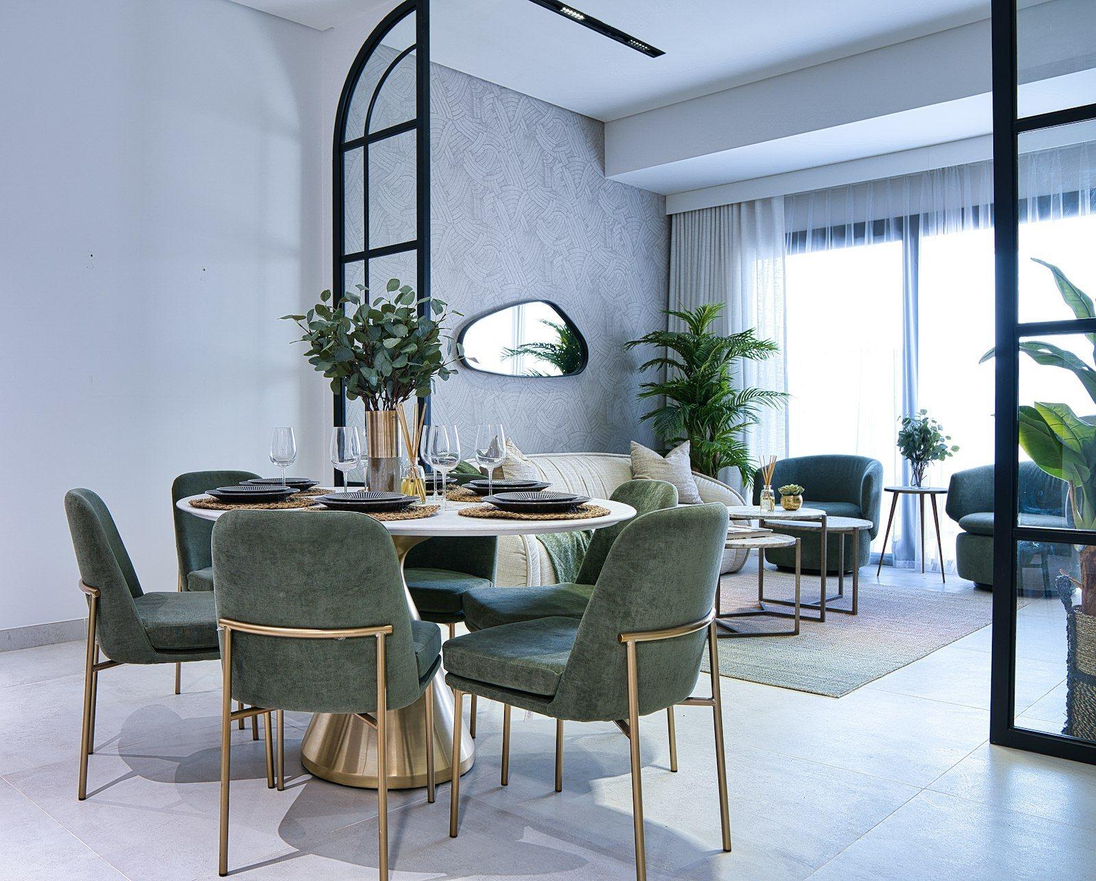 Dining area of the same reception: a round marble table on a brass base with six green velvet chairs, the arched steel screen and the living room beyond.