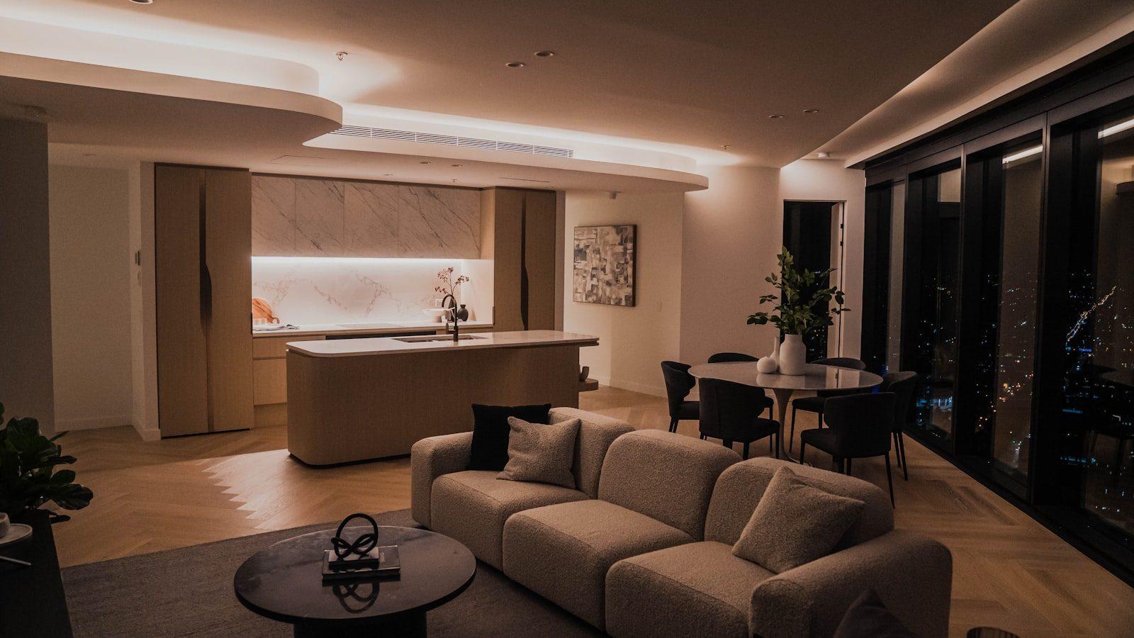 Evening in a high-floor reception room: herringbone timber flooring, an oak and marble island kitchen with a lit splashback, a bouclé sofa and round marble dining table, and a full-height sliding glazed wall onto a terrace with the city lights below.