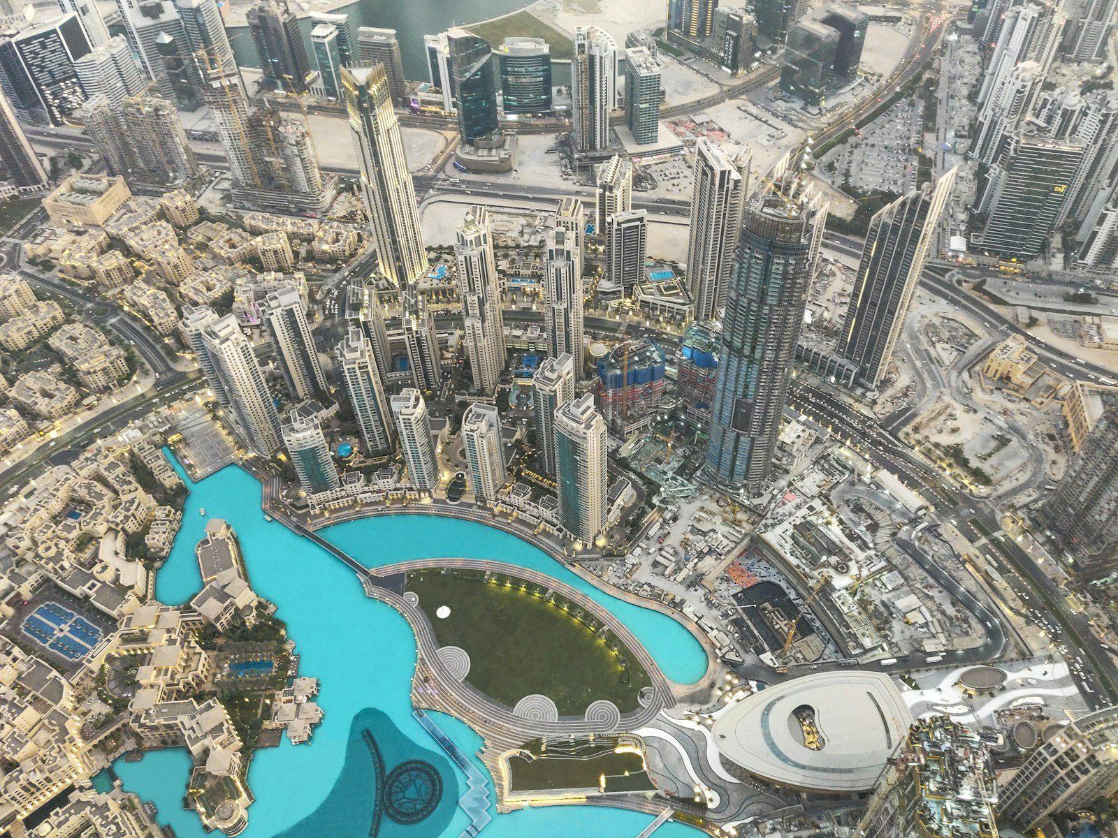 Downtown Dubai from above at dusk: the turquoise Burj Lake and the Dubai Fountain basin, the towers of the Opera District, Souk Al Bahar and the curved roof of Dubai Opera.