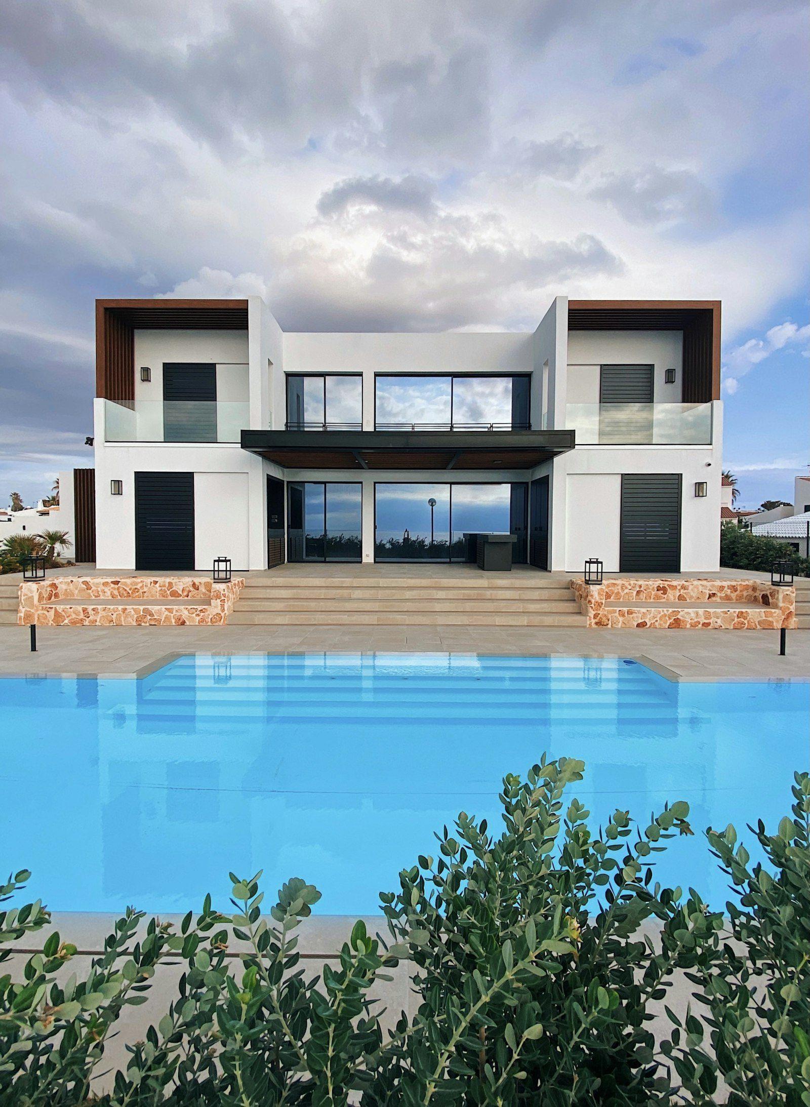 Contemporary white villa with a symmetrical elevation: a double-height glazed centre bay set between timber-lined recesses and stone-clad plinths, with a wide rectangular pool running the width of the terrace.