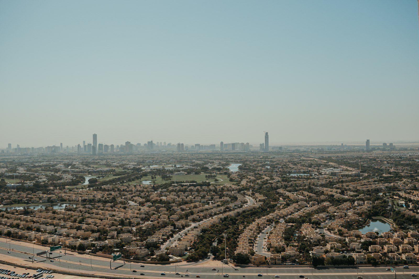 A Dubai villa estate seen from above: streets of low sand-coloured houses laid out around lakes and a green golf course, a multi-lane highway along the near edge and the towers of the city on the hazy horizon.