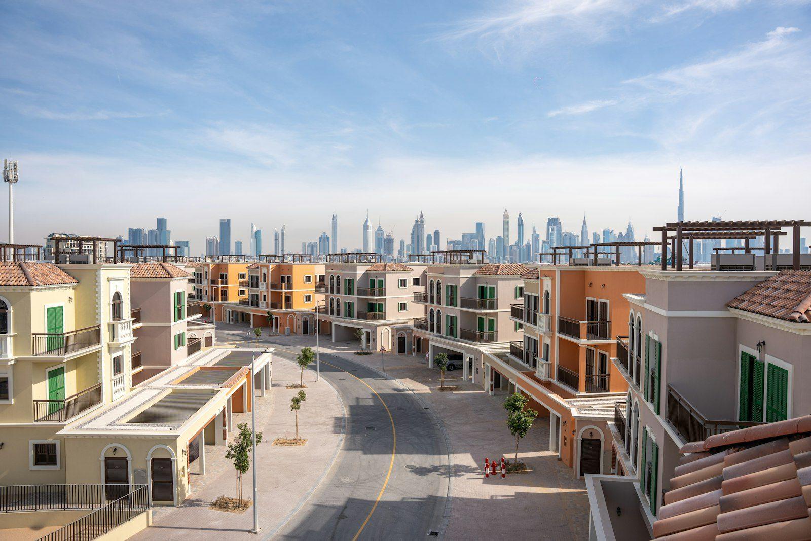 A Dubai townhouse community: terraces of cream houses with terracotta tile roofs and shuttered windows along a curving paved street, with the towers of Sheikh Zayed Road and the Burj Khalifa standing behind.