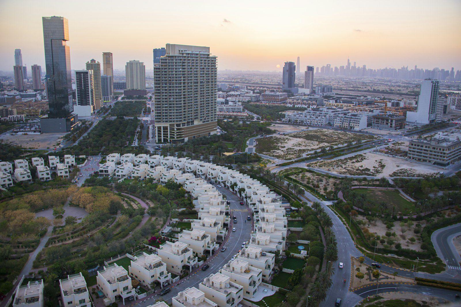 Jumeirah Village Circle at sunset from the air: white villas and townhouses curving around a landscaped district park, mid-rise towers behind them, undeveloped sand plots to one side and the city skyline on the horizon.