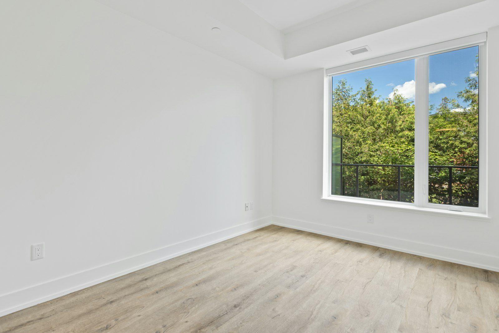 Unfurnished room in a newly finished house: white walls, pale oak flooring and a wide window with a railing looking into green trees.