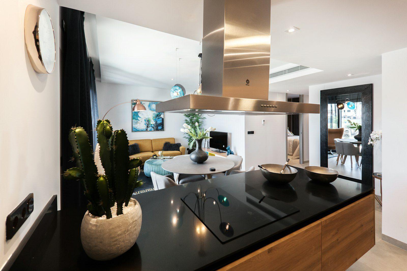 Furnished open-plan apartment: a black stone island with an induction hob under a brushed-brass extractor, oak drawer fronts, and beyond it a mustard sofa, round dining table and a bedroom doorway, with a ducted air-conditioning grille overhead.