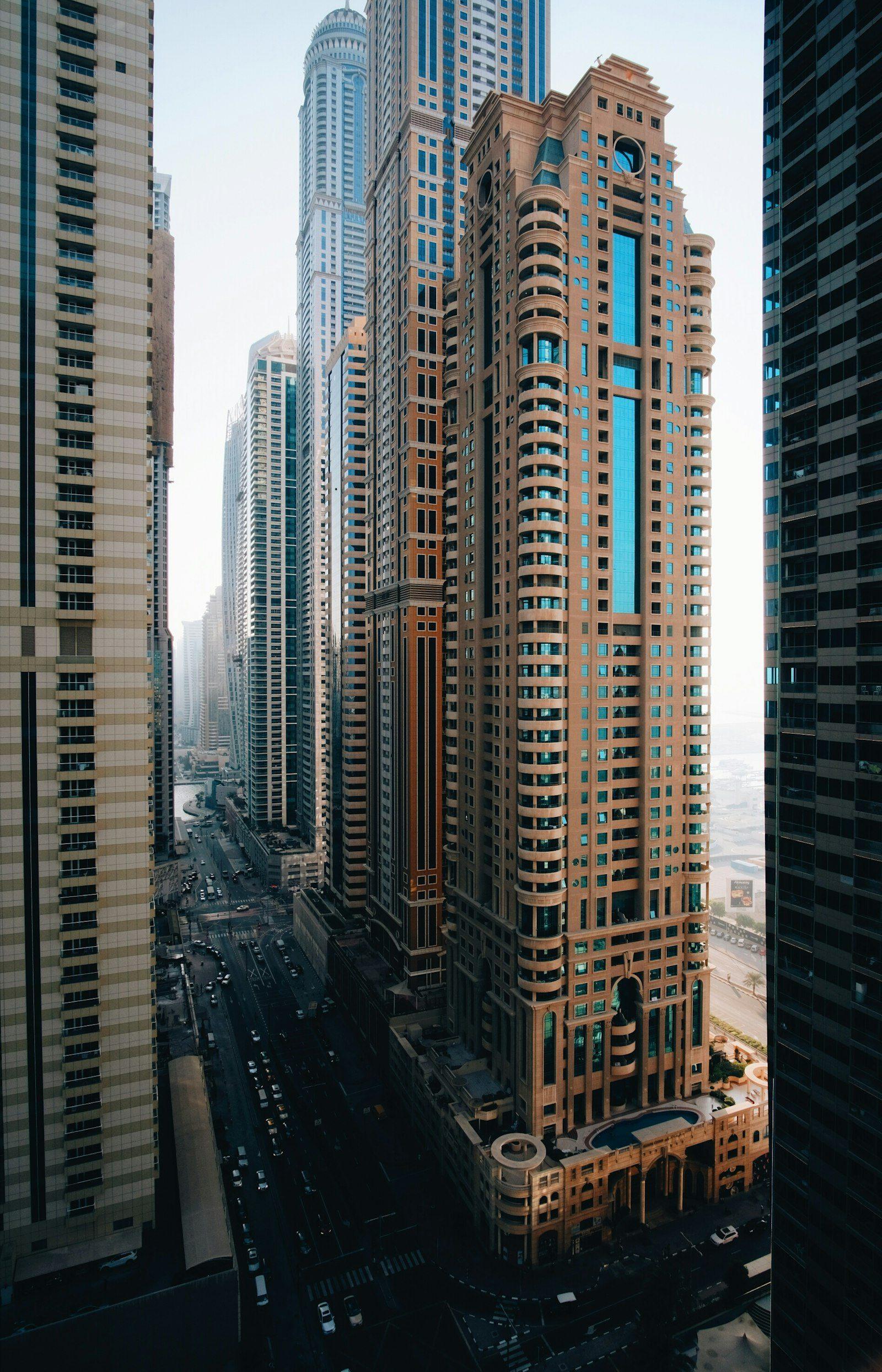 Dubai Marina from a high floor, looking down the canyon between the towers to Elite Residence and Princess Tower, traffic on the boulevard below and the sea haze to the right.