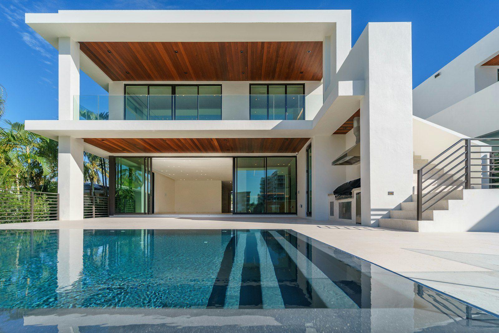 Contemporary white villa with deep cantilevered terraces and warm timber soffits, glass balustrades and full-height sliding walls open to the living room, an outdoor kitchen alongside and a long pool reflecting the house.
