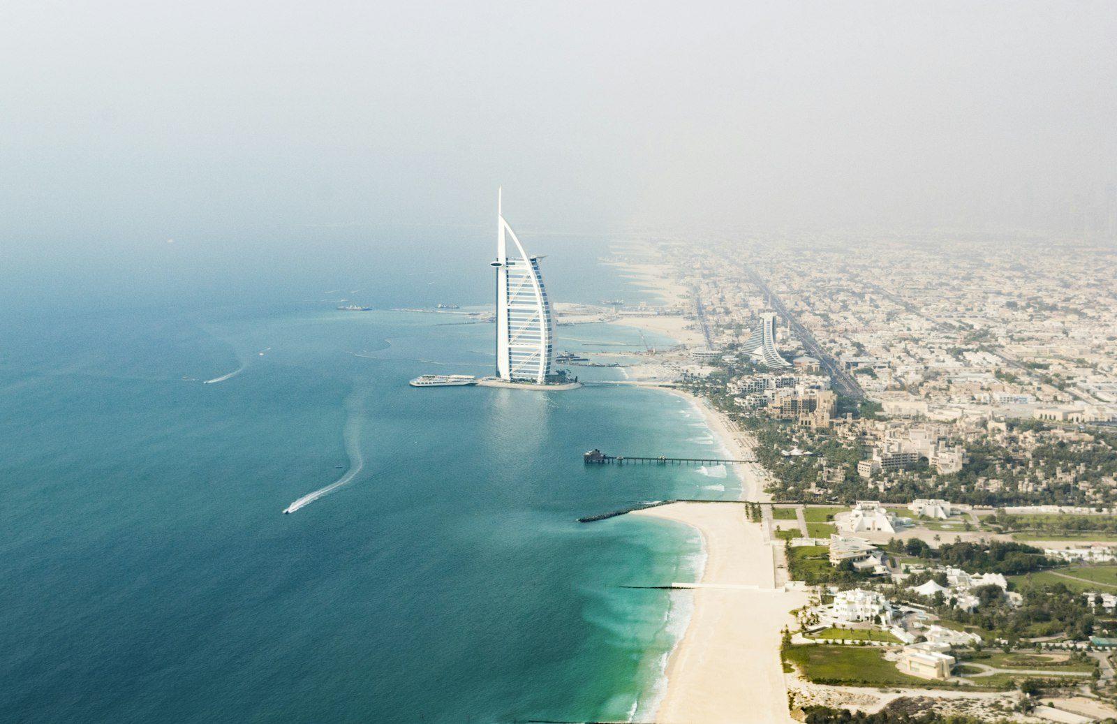 The Jumeirah coastline from the air: the Burj Al Arab on its island, turquoise shallows breaking along a wide sand beach, a pier and the low-rise resorts and city beyond.