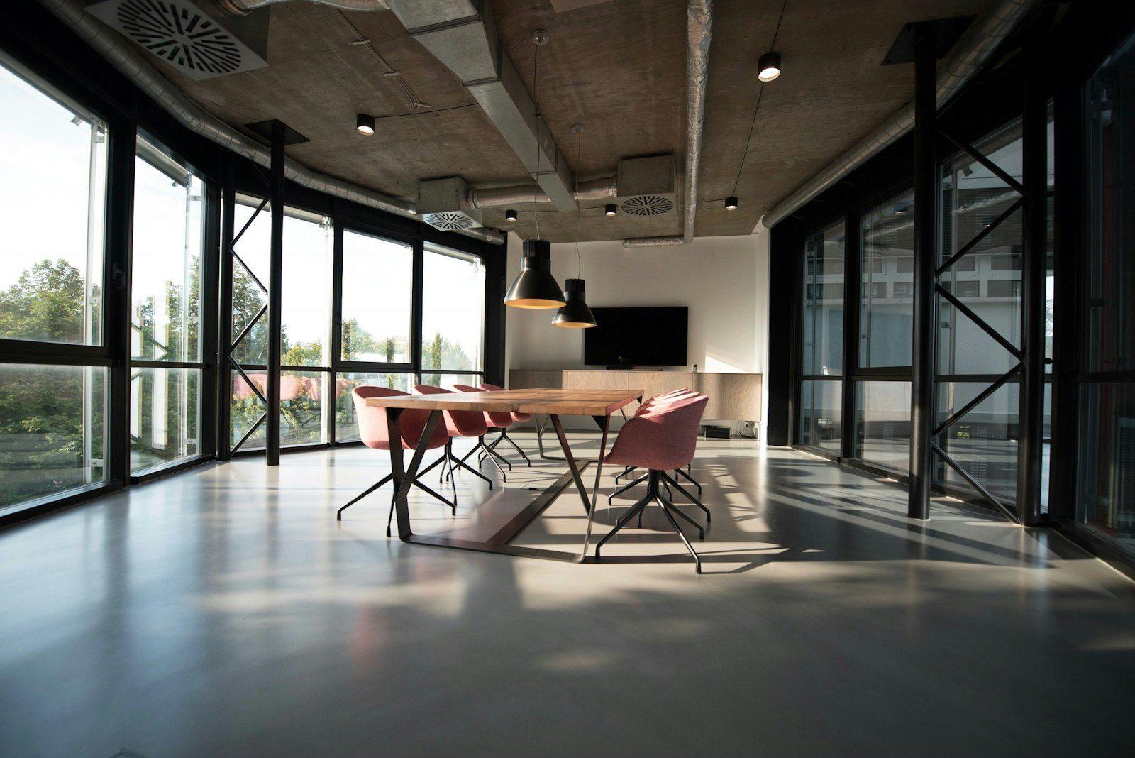 An empty modern office with floor-to-ceiling glazing, a long meeting table and black steel-framed glass partitions under an exposed concrete ceiling.