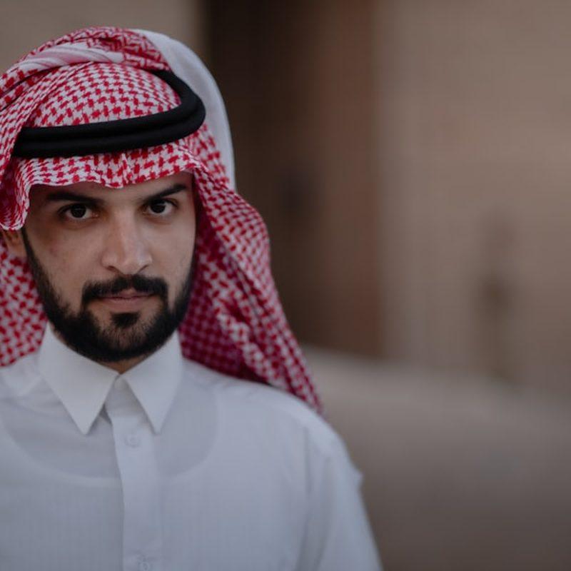 Portrait of a man in a white kandura and a red-and-white patterned ghutra against a softly blurred background.
