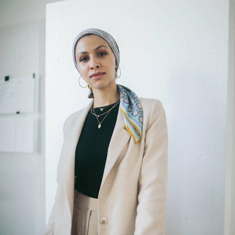 Portrait of a woman in a cream blazer and patterned headscarf standing against a plain white wall.