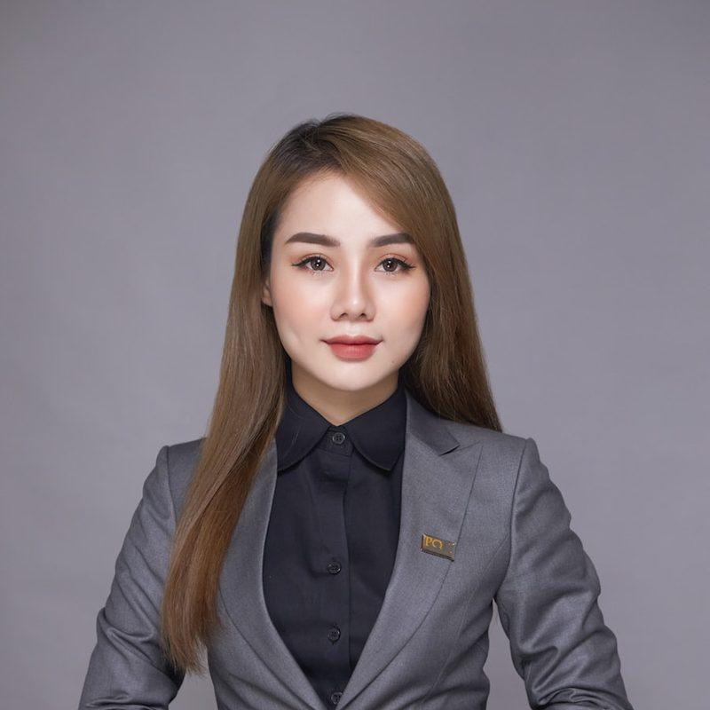 Portrait of a woman in a tailored grey suit, hands clasped, against a plain studio backdrop.