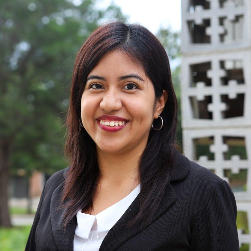 Portrait of a smiling woman in a black blazer and white top, photographed outdoors against a pale screen wall.
